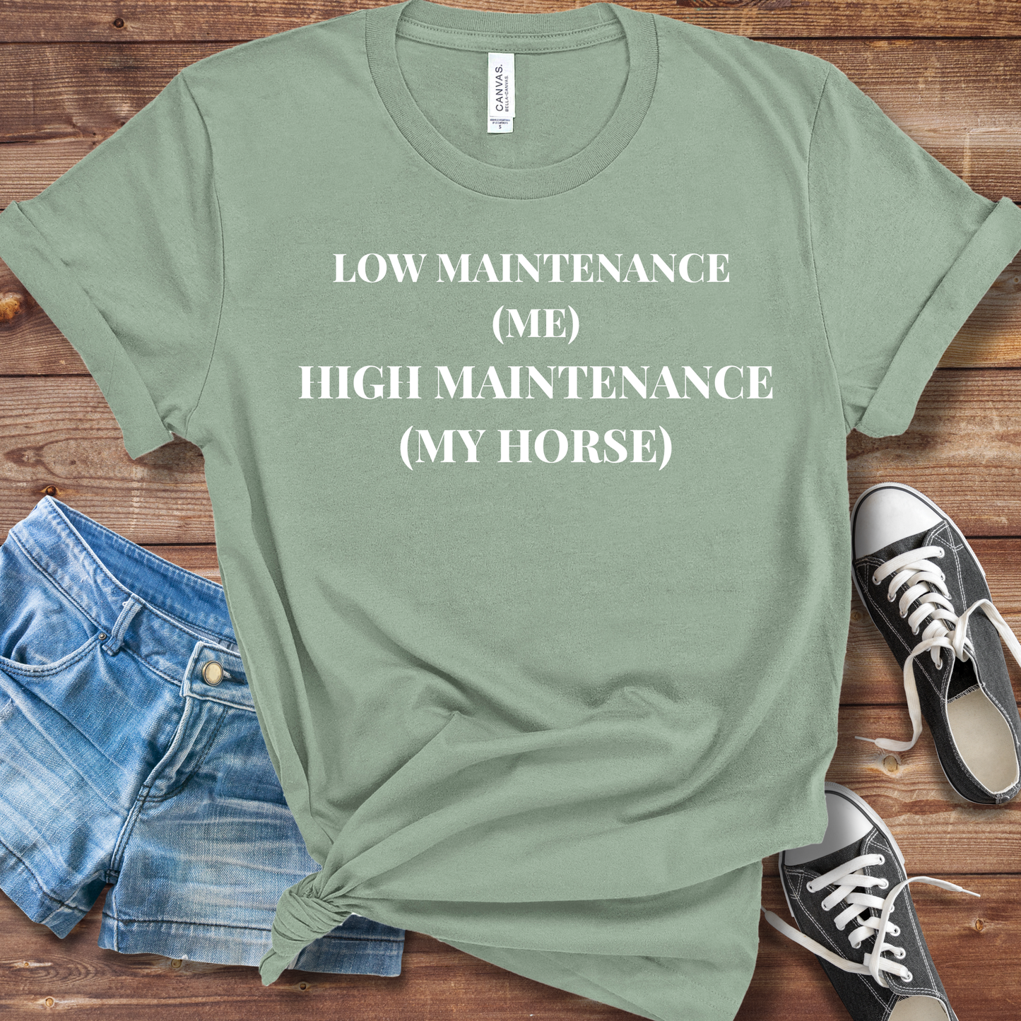 Green t-shirt with text 'LOW MAINTENANCE (ME) HIGH MAINTENANCE (MY HORSE)' on a wooden surface with shorts and sneakers.