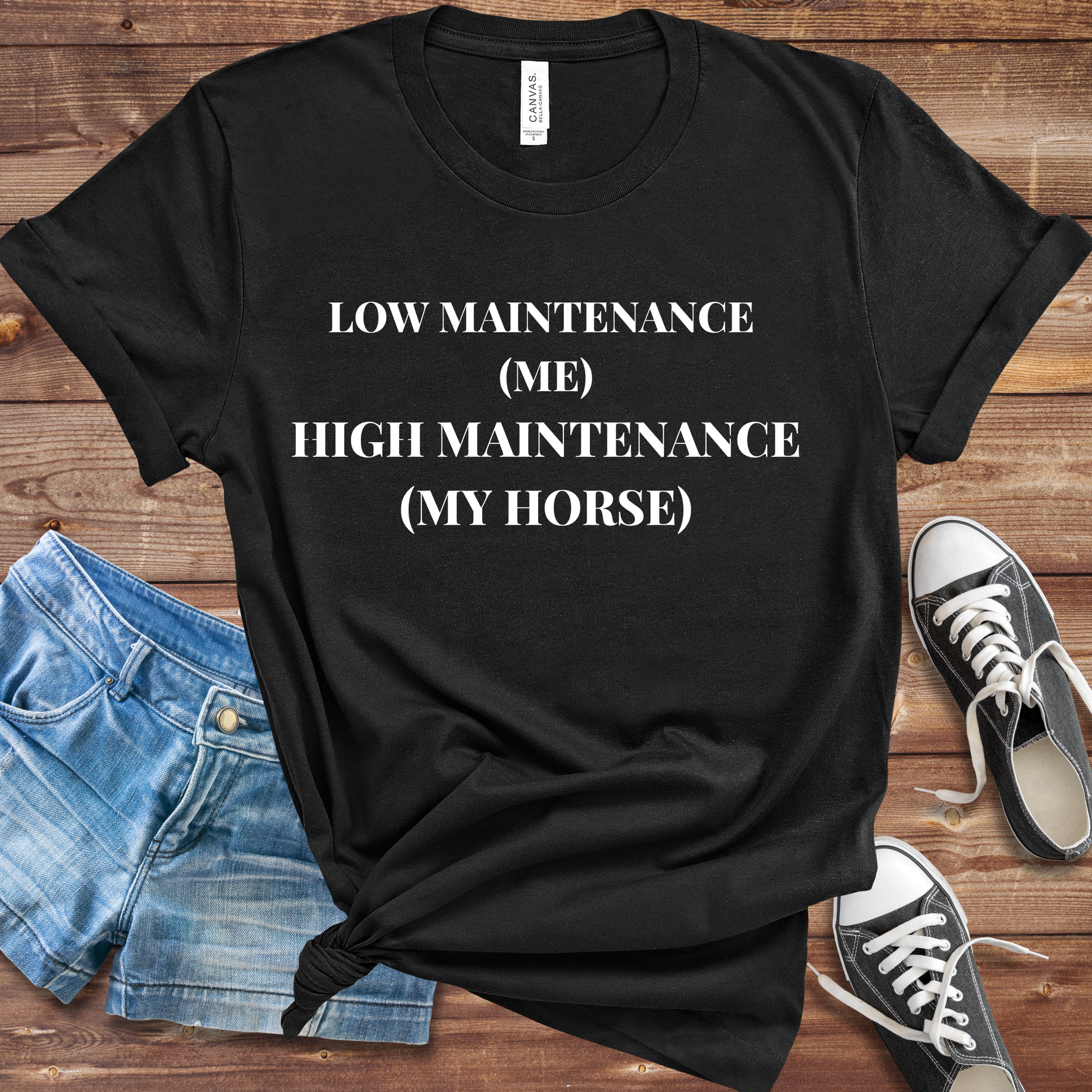 Black t-shirt with 'Low Maintenance (Me) High Maintenance (My Horse)' text on a wooden surface with shorts and sneakers.