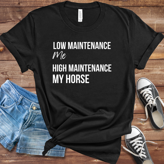 A black graphic t-shirt with white text reading 'LOW MAINTENANCE Me HIGH MAINTENANCE MY HORSE' is laid flat on a rustic wooden background, styled with blue denim shorts and black canvas sneakers.